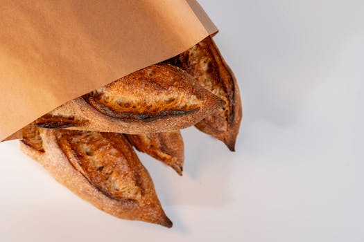 Fresh artisanal breads peeking out of a brown paper bag on white backdrop.